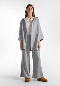 Grijze, losse capuchonjas over een wit, button-up shirt en grijze, wijde broek; inclusief zakken en bruine instapschoenen.