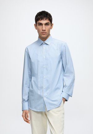 Jeune homme portant une chemise rayée bleu pâle à boutons et un pantalon crème, debout devant un fond blanc uni.