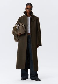 Olivengrønn ullfrakk med høy krage, med store knapper og rett snitt. Kombinert med mørke denimbukser og en beige veske.