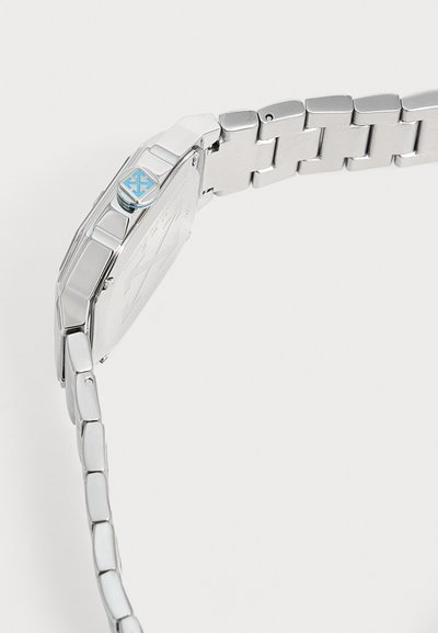 Bracelet de montre en métal argenté et vue latérale du boîtier montrant le logo bleu sur la couronne et des maillons métalliques détaillés sur un fond clair.