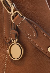 Angle d'un sac à main en cuir marron montrant des détails de couture, des accessoires en or et une étiquette ronde attachée par une sangle. Surface texturée visible.
