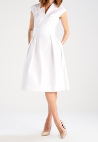 Vestido midi blanco con mangas cortas, escote en V y cintura ajustada. Presenta pliegues y bolsillos laterales. Textura de tela suave.