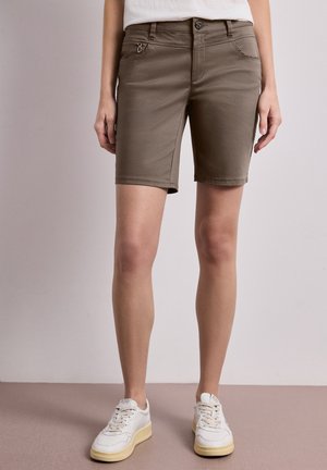 Kvinde iført olivengrønne knælange shorts og hvide sneakers, stående mod en ensfarvet neutral baggrund.