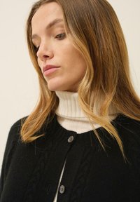 Svart stickad cardigan med kabeldesign, med rund halsringning och två mörka knappar, över en vit ribbad polotröja.