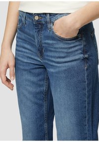 Vaqueros de mezclilla azul con un corte ajustado, que cuentan con cinco bolsillos, una cremallera y acentos de costura visibles. La tela parece suave y ligeramente texturizada.