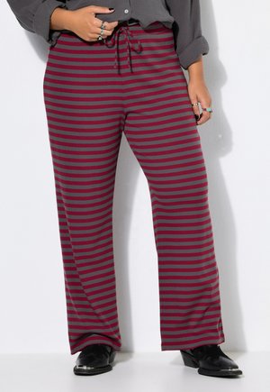 Personne portant un pantalon à jambes larges rayé horizontalement bordeaux et gris, des chaussures noires, une chemise grise et des bagues en argent aux mains.