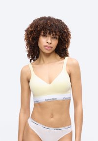 Kobieta o kręconych włosach, ubrana w jasnożółty biustonosz Calvin Klein i białą bieliznę Calvin Klein, stojąca na jednolitym tle.