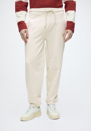 Personne portant un pantalon de jogging beige avec cordon, des baskets blanches et un haut à manches longues rouge et blanc, debout devant un fond uni.