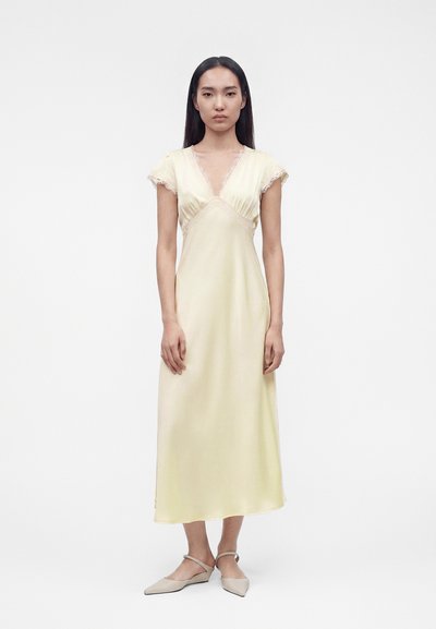 Woman with long dark hair wears a pale yellow satin midi dress with lace trim and beige pointed flats, standing against a plain white background.