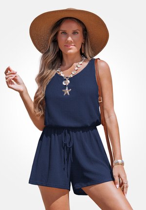 Granatowy kombinezon z teksturowanej tkaniny, bez rękawów, z elastycznym pasem i sznurkiem do wiązania. Uzupełniony naszyjnikiem z muszelek i kapeluszem z szerokim rondem.
