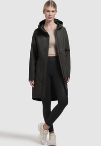 Lång svart vattentät kappa med huva, med en framknappstängning och sidofickor. Bärs över svarta leggings och sneakers.