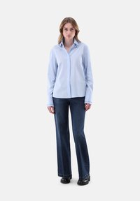 Donna in piedi di fronte, che indossa una camicia azzurro chiaro con bottoni, jeans dritti blu scuro e scarpe nere su uno sfondo bianco.