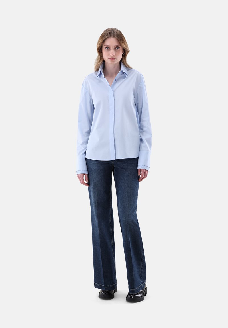Donna in piedi di fronte, che indossa una camicia azzurro chiaro con bottoni, jeans dritti blu scuro e scarpe nere su uno sfondo bianco.