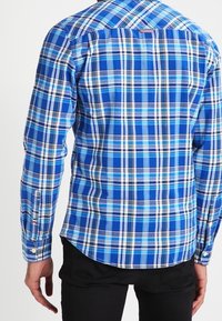 Uomo che indossa una camicia a maniche lunghe a quadri blu e bianchi e pantaloni neri, visto da dietro su uno sfondo semplice.