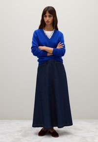 Blauwe gebreide V-hals trui met lange mouwen, gecombineerd met een lange donkere spijkerrok. Bruine instap-schoenen maken de outfit compleet.