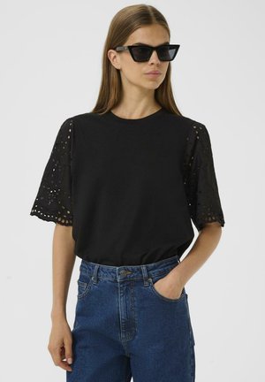 Zwarte t-shirt met ingewikkelde, laser-geknipte kantmouwen; comfortabele pasvorm; gecombineerd met hoge taille blauwe jeans; donkere zonnebril maakt de look af.