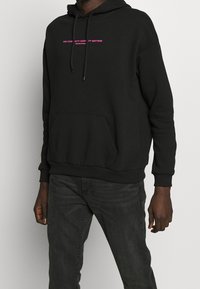 Svart huvtröja i bomullsblandning, med en framficka och dragsnodd på huvan. Texten i rosa lyder "EN GEMENSKAP MÅNGFALD ÄR VIKTIGT."