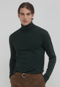 Maglione a collo alto di colore verde scuro realizzato in un tessuto liscio e elastico. Maniche lunghe, design aderente e dettagli a costine sul colletto.