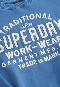 Felpa blu con testo ricamato bianco "SUPERDRY WORK-WEAR" e altri dettagli; include un colletto a coste e una texture morbida.