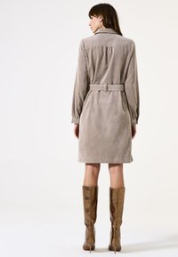 Vestido de pana marrón con cinturón en la cintura, mangas largas y yugo en la parte posterior, combinado con botas doradas brillantes hasta la rodilla. Textura suave y diseño ajustado.