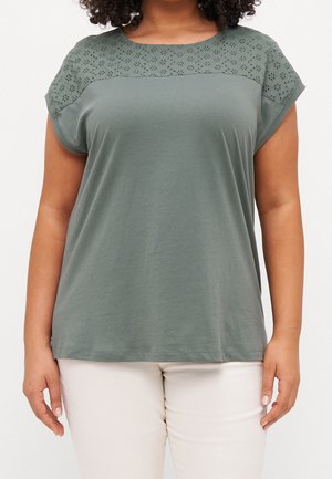 Vrouw draagt een groen shirt met korte mouwen en bloemenoogjesdetail op de schouders en witte broek, staand met ontspannen handen langs het lichaam.