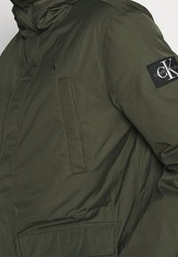 Olivově zelená podšívková bunda s vysokým límcem, zapínáním na zip, boční kapsou a �černou nášivkou s logem Calvin Klein na rukávu.