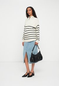 Femme portant un pull rayé blanc et noir, une jupe midi bleu clair avec une fente à l'avant, des chaussures à talons noires, tenant un sac à main noir contre un fond blanc.