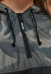 Chaqueta con capucha camuflada y cremallera media en el frente, hecha de una tela ligera. Presenta detalles en negro, cordones y una etiqueta con el logotipo en el cuello.