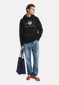 Felpa nera con logo, jeans blu, sneakers arancioni, che tiene una borsa a tracolla blu navy. Outfit casual adatto per l'uso quotidiano.