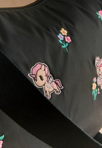 Mustast kangast tikitud värvilise ükssarviku ja lilledega, kujutatud on ka tugev must rihm, mis on kaldus pinnale asetatud.