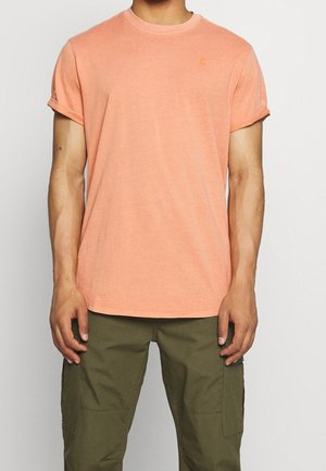 Homme portant un t-shirt manches courtes couleur saumon avec les manches retroussées et un pantalon cargo vert olive, sur un fond uni.