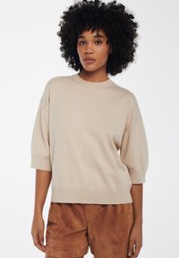 Beige gebreide trui met korte, brede mouwen en een geribbelde zoom. Draagt over bruine suède shorts. Soepele textuur, casual pasvorm.