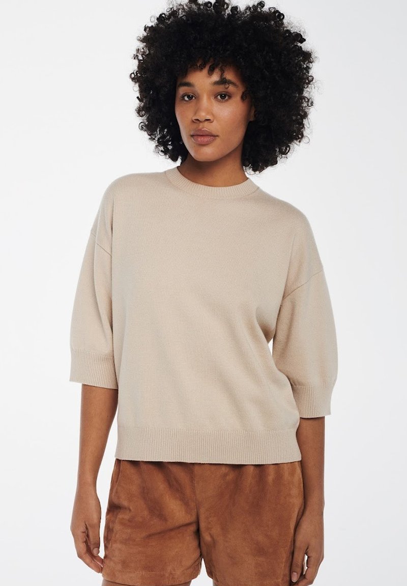 Beige gebreide trui met korte, brede mouwen en een geribbelde zoom. Draagt over bruine suède shorts. Soepele textuur, casual pasvorm.