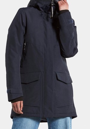 Frau trägt eine schwarze Didriksons Winterjacke mit Kapuze, Frontreißverschluss, Klappentaschen und verstellbaren Handgelenkriemen über blauer Jeans.