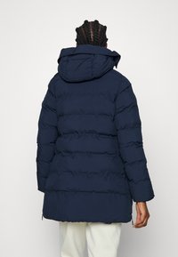 Granatowy pikowany płaszcz z kapturem, z poziomymi przeszyciami i bocznymi zamkami, wykonany z gładkiego, odpornego na warunki pogodowe materiału.