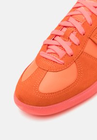Zapatilla naranja con una mezcla de texturas lisas y de ante, con cordones de color rosa brillante y una suela de goma. Se pueden ver detalles de costura destacados.