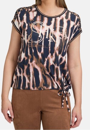 Vrouw draagt een top met korte mouwen met dierenprint en de tekst "SHINE" en een zijstrik, gecombineerd met een bruine broek.