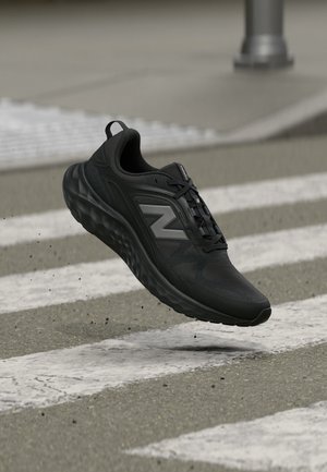 Chaussure de course New Balance noire avec une semelle épaisse, volant au-dessus d'un passage piéton sur une route asphaltée.