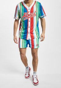 Monivärinen raitainen lyhythihainen nappipaita ja siihen sopivat shortsit sinisen, vihreän, punaisen ja keltaisen sävyissä, joissa logo rinnassa ja helmoissa.