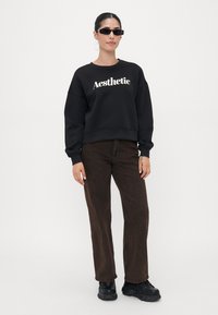 Felpa nera con la scritta "Aesthetic" in caratteri bianchi, abbinata a jeans dritti marroni e sneakers nere chunky. Presenta occhiali da sole e capelli lisci.
