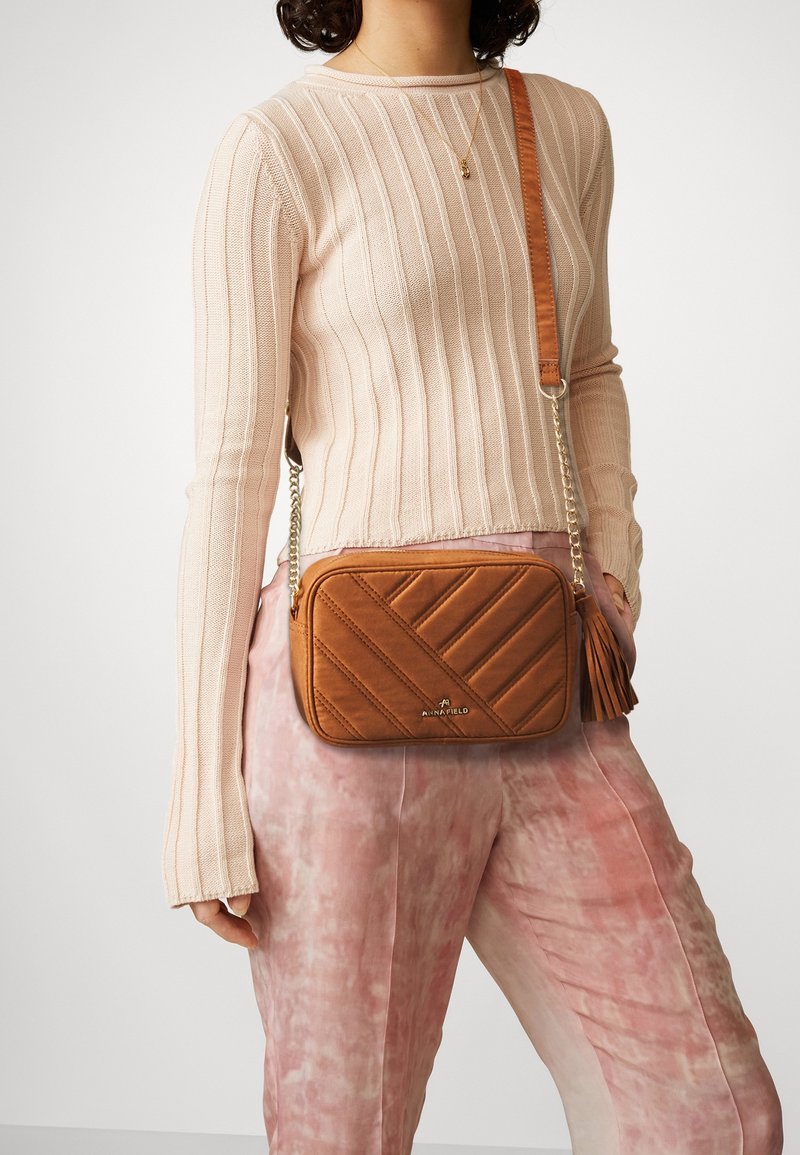 Brun quiltad crossbody-väska med en tofsdetalj; tillverkad av konstläder. Har en kort kedjesnurra och en mjuk yta.