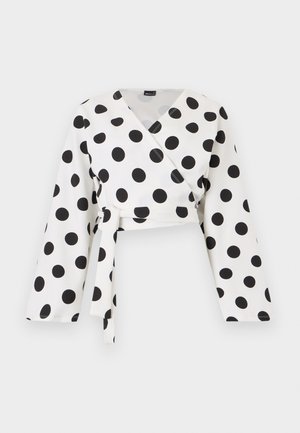 Witte blouse met lange mouwen, grote zwarte polkadots en een vooraan gestrikte riem, afgebeeld op een effen witte achtergrond.