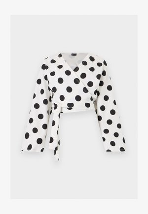 Witte blouse met lange mouwen, grote zwarte polkadots en een vooraan gestrikte riem, afgebeeld op een effen witte achtergrond.
