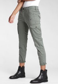 Pantaloni cargo verde oliva con gambe affusolate, multiple tasche e orli arrotolati, abbinati a stivaletti neri in pelle.