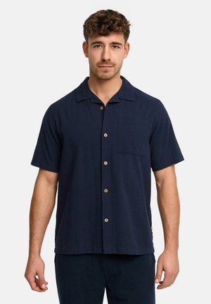 Mand med kort krøllet hår iført en mørk marineblå kortærmet skjorte med knapper og brystlomme, stående foran en ensfarvet lys baggrund.