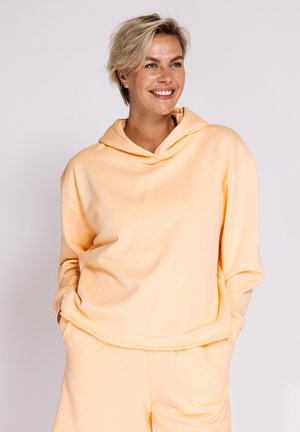 Smiling person with short blonde hair wearing a loose peach hoodie and matching pants, hands in pockets against a plain background.
