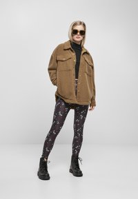 Brun fleecejakke med to brystlommer, svart høyhalset genser, blomstrete leggings i mørke farger, og snørebånd sorte støvler.