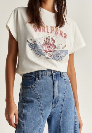 T-shirt en coton blanc avec un motif graphique d'ailes et de flammes. Associé à un jean en denim bleu clair taille haute à coupe décontractée.