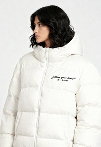 Doudoune blanche avec col montant, fermeture éclair, texte brodé "suis ton cœur" et motif matelassé pour l'isolation.