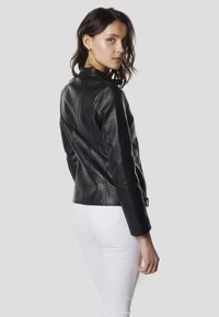 Čierna kožená bunda s priliehavým strihom, stojatým golierom a panelovým zdobením. Kombinovaná s bielymi úzkymi džínsami.